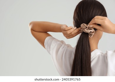 Young Asian woman doing ponytail with scrunchy on light grey background, back view