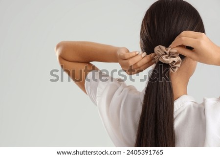 Young Asian woman doing ponytail with scrunchy on light grey background, back view