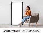 Young Asian woman drinking coffee, having break, relaxing in armchair near giant cellphone with empty screen against white studio wall, mockup for cool new mobile app or online ad design