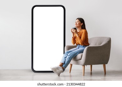 Young Asian Woman Drinking Coffee, Having Break, Relaxing In Armchair Near Giant Cellphone With Empty Screen Against White Studio Wall, Mockup For Cool New Mobile App Or Online Ad Design