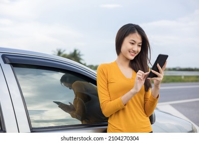 A young Asian woman drives a beautiful nature drive. she was standing in front of the car on the roadside. she uses the smartphone to call services. - Powered by Shutterstock