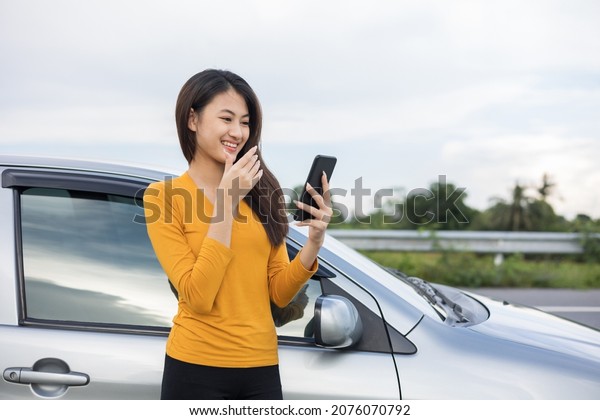 A young Asian woman drives a beautiful\
nature drive. she was standing in front of the car on the roadside.\
she uses the smartphone to call\
services.