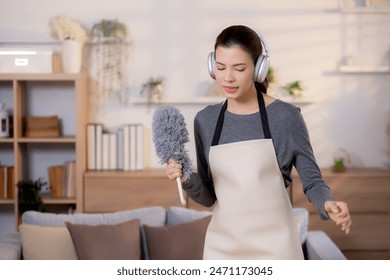 Young asian woman dusting while listening to music with headphones in a bright, organized living room, maid doing housework enjoying music and sing while dusting at home, lifestyle concept. - Powered by Shutterstock