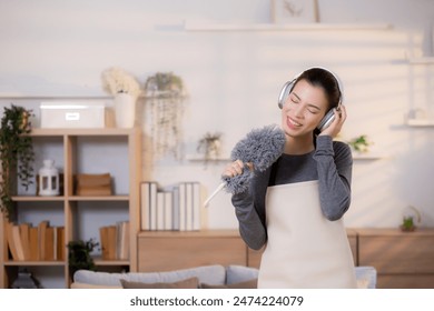Young asian woman dusting while listening to music with headphones in a bright, organized living room, maid doing housework enjoying music and sing while dusting at home, lifestyle concept. - Powered by Shutterstock