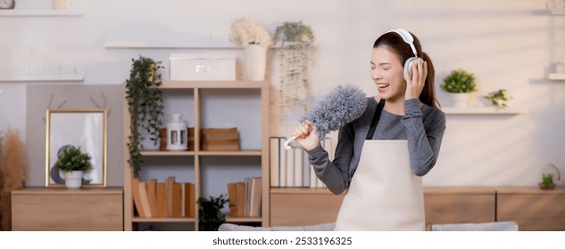 Young asian woman dusting while listening to music with headphones in a bright, organized living room, maid doing housework enjoying music and sing while dusting at home, lifestyle concept. - Powered by Shutterstock