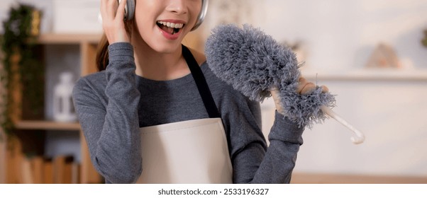 Young asian woman dusting while listening to music with headphones in a bright, organized living room, maid doing housework enjoying music and sing while dusting at home, lifestyle concept. - Powered by Shutterstock