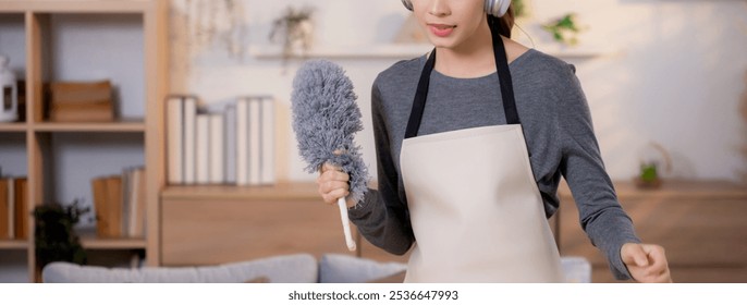 Young asian woman dusting while listening to music with headphones in a bright, organized living room, maid doing housework enjoying music and sing while dusting at home, lifestyle concept. - Powered by Shutterstock