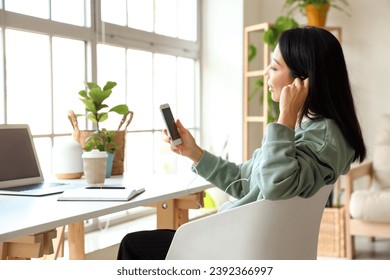 Young Asian woman with earphones and mobile phone at home office - Shutterstock ID 2392366997