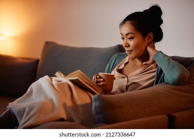 Young Asian woman enjoying in cup of tea while relaxing on the sofa and reading a book at home.  - Powered by Shutterstock