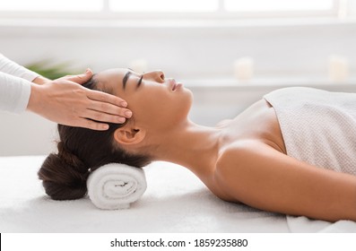 Young Asian Woman Enjoying Relaxing Head Massage During Beauty Treatment In Spa Salon, Having Wellness Day At Cosmetology Cabinet, Lying On Table With Rolled Towel Under Head, Side View