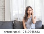 Young asian woman experiencing a headache sitting on a sofa with a pained expression, serious mood, health, wellness, stress, headaches and everyday struggles, pain and importance of self-care at home