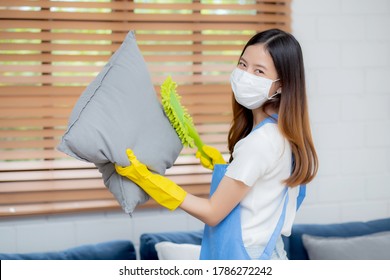 Young Asian Woman In Face Mask And Gloves Cleaning Dust With Duster On Sofa And Cushions At Home, Girl Doing Housework For Hygiene At House, Housekeeper And Service, Health Care And Lifestyle Concept.