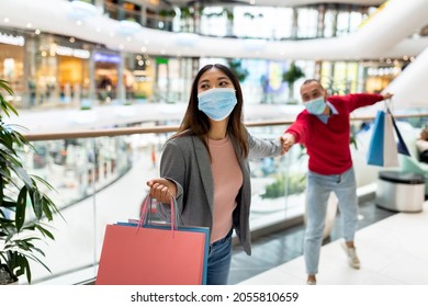 Young Asian woman in face mask holding shopper bags, pulling her unwilling boyfriend by hand at supermarket. Millennial multiracial couple shopping at huge mall. Shopaholism concept - Powered by Shutterstock