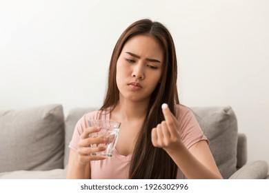 Young Asian Woman Feeling Unwell And Holding Glass Of Water And Taking Pill While Sitting On Sofa At Home. Asian Woman Stressed And Sick 