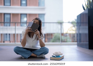 Young asian woman freelancer is hiding her face with a notebook and thinking about new ideas while relaxing on the terrace with tablet, book and headphones - Powered by Shutterstock