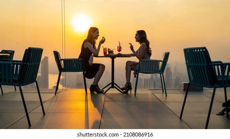 Young Asian Woman Friends Celebrating Dinner Party Meeting At Skyscraper Rooftop Restaurant Bar In Metropolis At Summer Sunset. Beautiful Girl Enjoy Outdoor Lifestyle Together On Holiday Vacation.