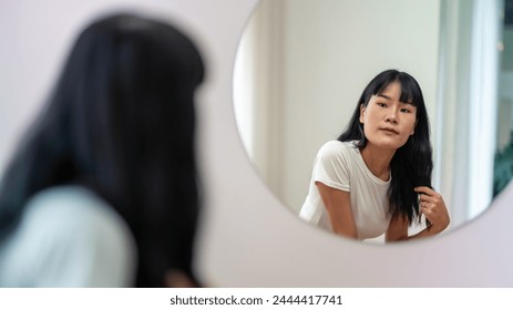 A young Asian woman gazes at her reflection in a round mirror in a bright, minimalist room. - Powered by Shutterstock