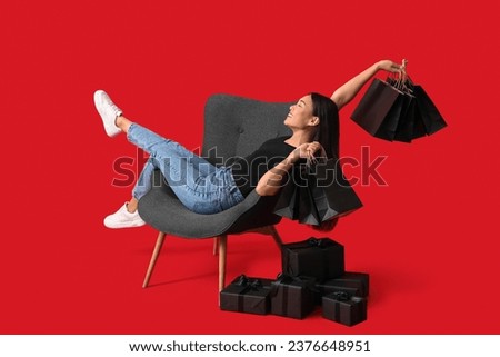 Young Asian woman with gift boxes and shopping bags sitting on chair against red background. Black Friday sale