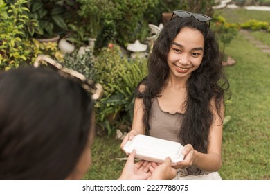 A young asian woman gives takeout food or dessert inside a styrofoam container to her friend. Lighthearted outdoor scene. - Shutterstock ID 2227788075