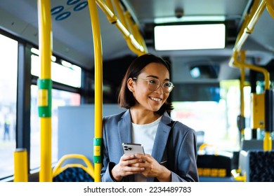 Young asian woman in glasses smiling and using cellphone while standing in a bus - Powered by Shutterstock