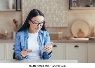 Young asian woman happily shopping online sitting at home in the kitchen, woman using smartphone and bank credit card for online shopping
