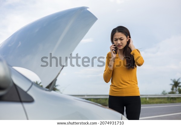 Young asian woman has\
broken down car on the road she feeling serious and stressed. Look\
for someone help. Using smartphone Call Emergency assistance or\
insurance service.