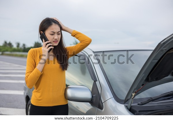 Young asian woman has\
broken down car on the road she feeling serious and stressed. Look\
for someone help. Using smartphone Call Emergency assistance or\
insurance service.