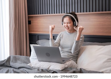 Young asian woman with headphone put laptop on pillow to working about business and raising hand when finish work while relaxing by listening to music and sitting on the bed in home bedroom
