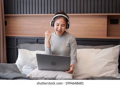 Young asian woman with headphone showing yes gesture with hand after receive email good news while sitting to working and listening to music from laptop on the bed in home bedroom
