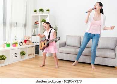 Young Asian Woman And Her Daughter Enjoying Singing And Playing Music In The Living Room.