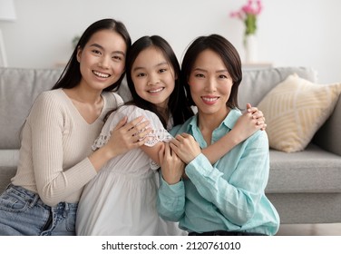 Young Asian woman with her mature mother and adorable little daughter hugging and looking at camera at home. Multi generation family smiling, expressing love and fondness