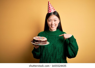 Young Asian Woman Holding Birthday Cake Wearing Party Hat Over Yellow Isolated Background With Surprise Face Pointing Finger To Himself