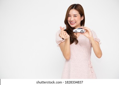 Young Asian Woman Holding Car Model And Showing Mini Heart Sign Isolated On White Background, Travel Insurance Concept