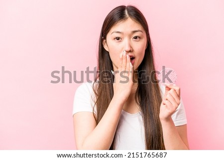 Young asian woman holding facial sponge isolated on pink background is saying a secret hot braking news and looking aside
