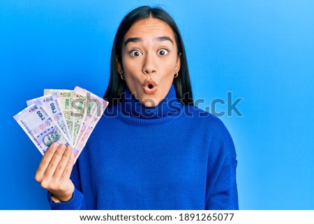 Young asian woman holding indian rupee banknotes scared and amazed with open mouth for surprise, disbelief face 