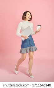 Young Asian Woman Holding Laptop And Coffee While Walking Over Pink Background.