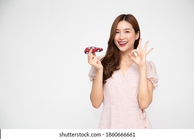 Young Asian Woman Holding Red Car Model And Showing Ok Sign Isolated On White Background, Travel Insurance Concept