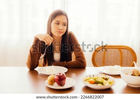 Young asian woman hungry but bored eating on table full of bland food sitting looking at food with boredom and having anorexia, not happy to eat and not having good appetite.