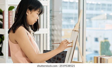 Young Asian woman intently paints on canvas in sunlit studio, surrounded by books and laptop - Powered by Shutterstock