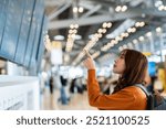 Young asian woman in international airport, using mobile smartphone and checking flight at the flight information board