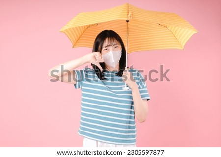 Young asian woman isolated on pink background showing thumb down wears protective mask to reduce breathing pollutants, hides under umbrella. Air pollution concept