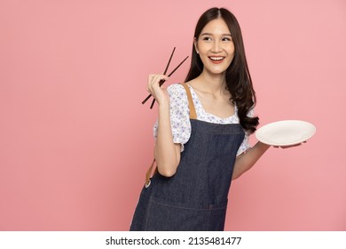 Young Asian woman with jeans apron holding chopsticks and empty white plate or dish isolated on pink background