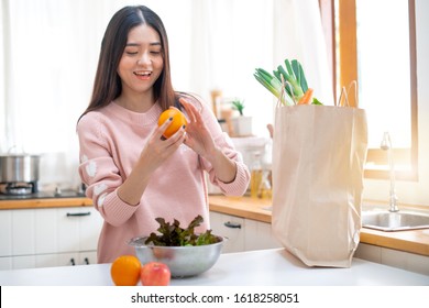 Young Asian woman in the kitchen holding orange, grocery shopping bag on table - Shutterstock ID 1618258051
