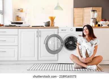 Young Asian Woman With Laundry Basket Sitting Near Washing Machine In Kitchen