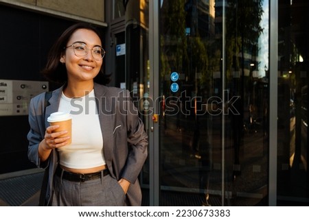 Young asian woman leaving the office building holding takeaway coffee and smiling