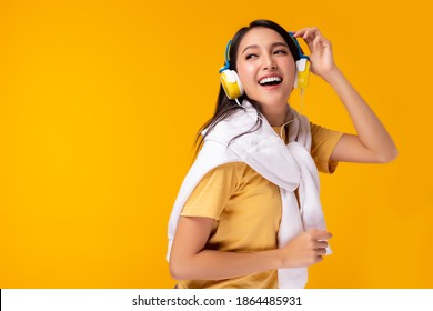 Young Asian Woman Listening Music In Headphones And Singing Hanging Sweater On Shoulder  On Yellow Background. Charming Girl Dancing. Attractive Lady Enjoying Favorite Song. Copy Space