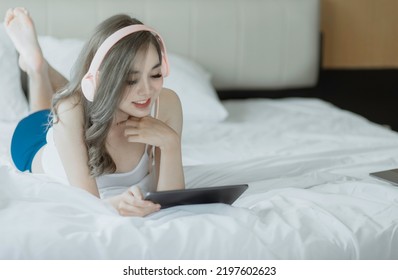 Young Asian Woman Listening To Music From Digital Tablet And Relaxing While Sitting On Bed At Home.