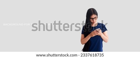 Young Asian woman with long hair wearing eyeglasses standing and playing on her phone, gray background with space for text caption