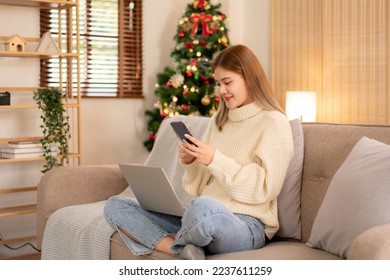 Young asian woman looking business data on smartphone and working on laptop while wearing sweater and sitting to working in holiday on sofa in cozy living room of christmas decoration at home. - Powered by Shutterstock