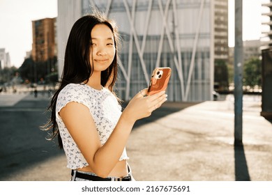 Young Asian Woman Looking At Camera While Using Her Mobile Phone Outdoors On The Street.